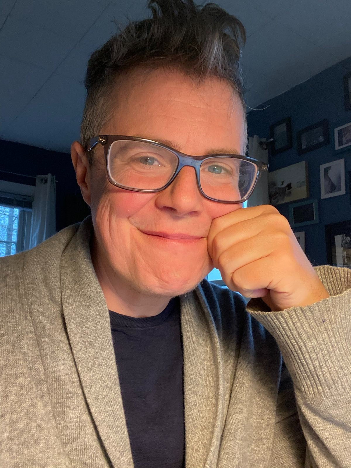 Image description: bespectacled, smiling white trans masc with grey fauxhawk in blue shirt and grey cardigan. 